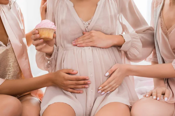 Обрезанный вид подруг, касающихся живота молодой беременной женщины, держащей розовый кекс на детской вечеринке — стоковое фото