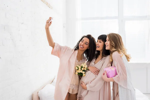 Sonrientes novias multiculturales y feliz mujer embarazada sosteniendo flores y presente mientras toma selfie en baby shower - foto de stock