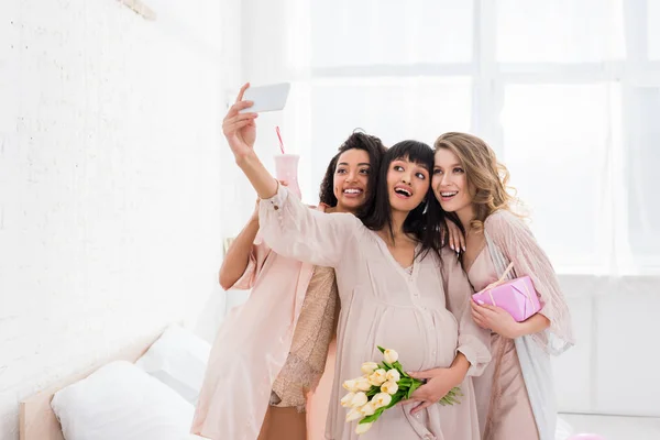Novias multiétnicas y feliz mujer embarazada sosteniendo batido, flores y regalo mientras toma selfie en baby shower - foto de stock