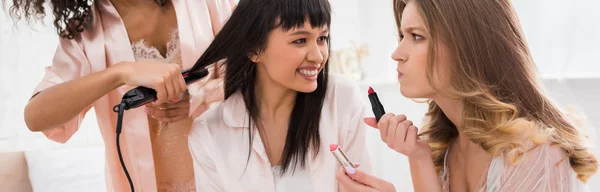 Panoramic shot of smiling multicultural girlfriends doing hair styling and makeup with lipsticks on bachelorette party — Stock Photo