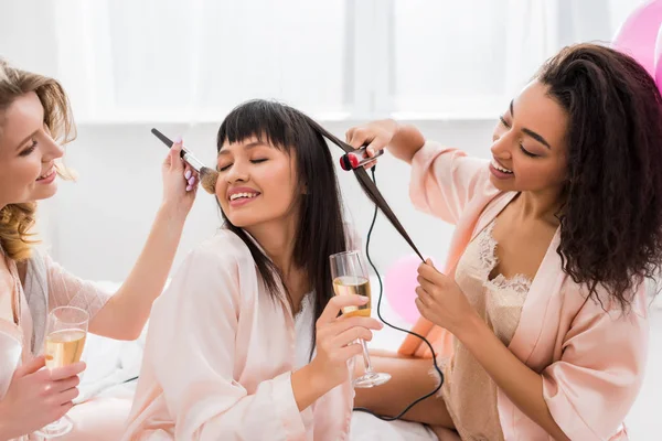 Feliz multiétnica novias haciendo peinado, maquillaje y beber champán en despedida de soltera - foto de stock