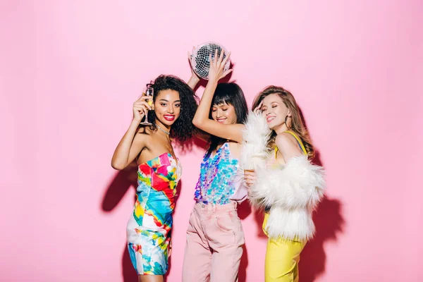 Happy multicultural girlfriends with champagne and disco ball dancing on pink — Stock Photo