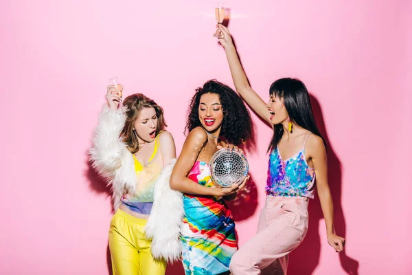 Excited multicultural girlfriends with champagne and disco ball having fun on pink — Stock Photo