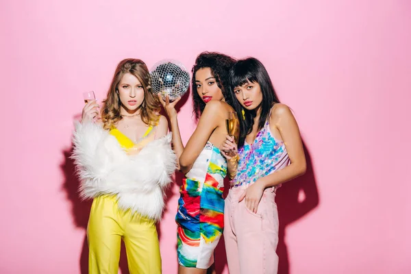 Emotional multiethnic girlfriends with champagne and disco ball posing on pink — Stock Photo