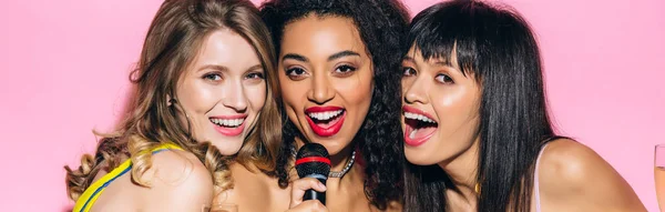 Plano panorámico de chicas multiétnicas felices atractivas cantando karaoke con micrófono, aislado en rosa - foto de stock