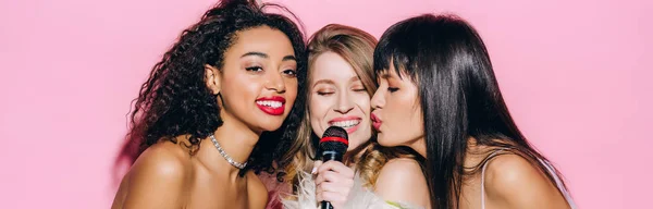 Panoramic shot of cheerful multicultural girls singing karaoke with microphone, isolated on pink — Stock Photo