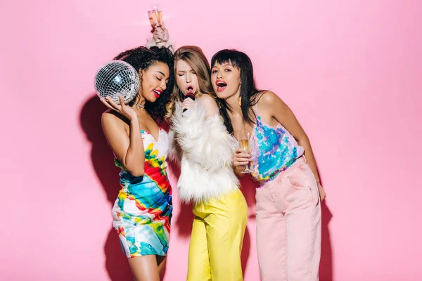 Emotional multiethnic girls holding champagne glasses and disco ball while singing with microphone on pink — Stock Photo