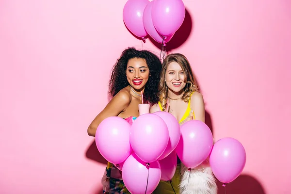 Multicultural smiling girlfriends holding milkshakes on pink with balloons — Stock Photo