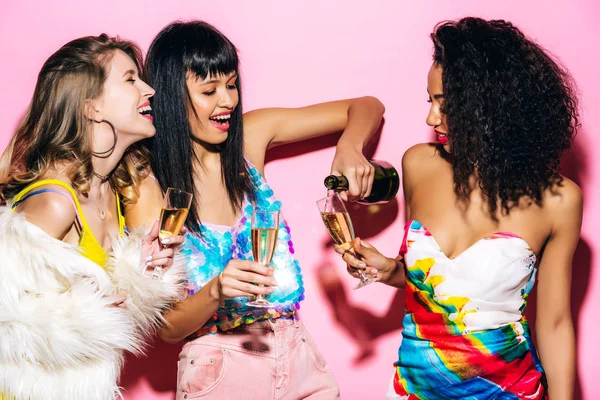 Emotionale multikulturelle Freundinnen gießen Champagner aus der Flasche in Gläser auf rosa — Stockfoto