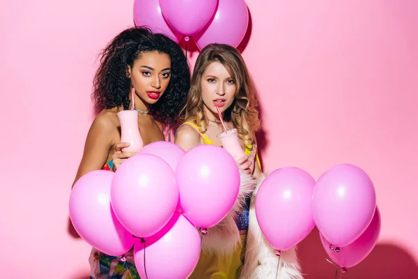 Sexy multikulturelle Mädchen trinken Milchshakes auf rosa mit Luftballons — Stockfoto