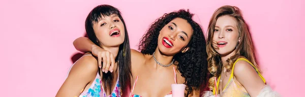 Panoramic shot of happy fashionable multiethnic girls holding glasses with milkshakes on pink — Stock Photo