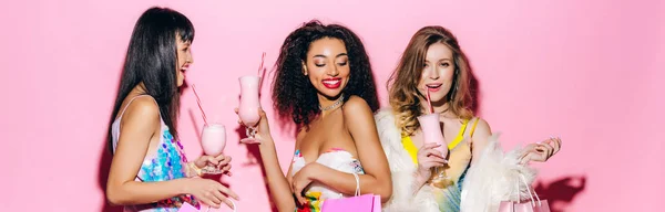 Panoramic shot of cheerful stylish multiethnic girlfriends drinking milkshakes on pink — Stock Photo