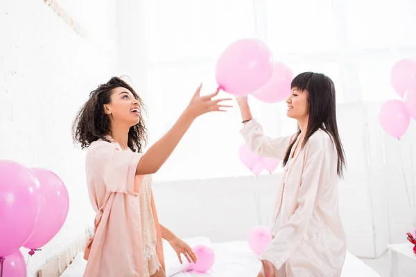 Chicas emocionales multiculturales en albornoces sosteniendo globos rosados en despedida de soltera - foto de stock