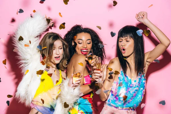 Eccitate ragazze multiculturali alla moda che ballano con bicchieri di champagne su rosa con coriandoli — Foto stock