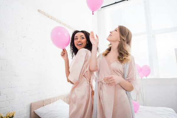 Happy multiethnic girls in bathrobes holding pink balloons on bachelorette party — Stock Photo