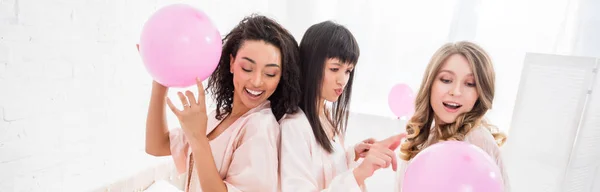 Panoramic shot of multicultural girls having fun with pink balloons in bedroom — Stock Photo