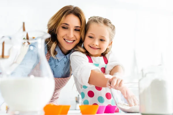 Selective focus of beautiful mother and daughter preparing cupcakes with ingredients including milk and flour in kitchen — Stock Photo