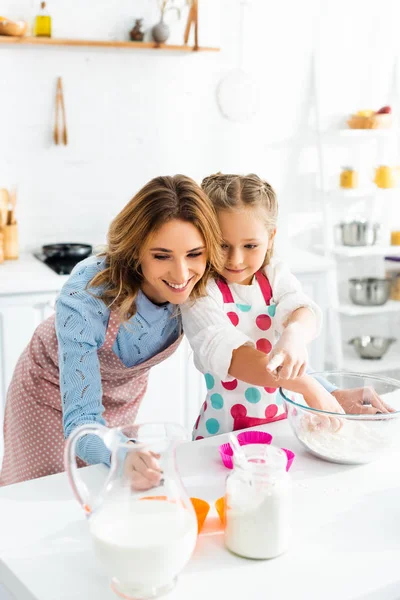 Attractive mother and cute daughter preparing cupcakes with ingredients including milk, flour in kitchen — Stock Photo