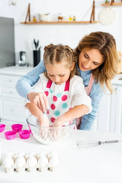 Hermosa madre añadiendo harina mientras linda hija amasando masa en un tazón - foto de stock