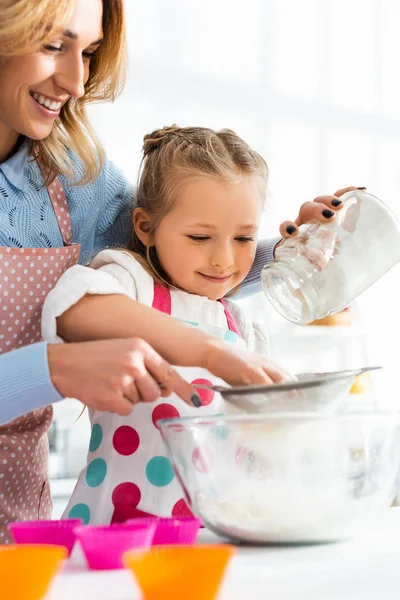 Selective focus of beautiful mother and cute daughter sifting flour through a sieve into bowl — Stock Photo