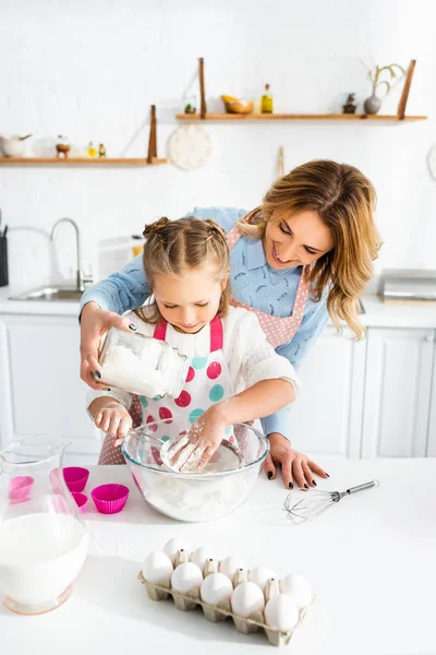 Schöne Mutter und niedliche Tochter sieben Mehl in eine Schüssel auf dem Tisch in der Nähe von Teigformen, Eiern, Milchkrug und Luftballonbesen — Stockfoto