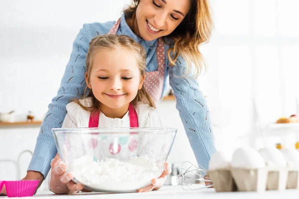 Selective focus of smiling mother and daughter near bowl with flour at kitchen table — Stock Photo