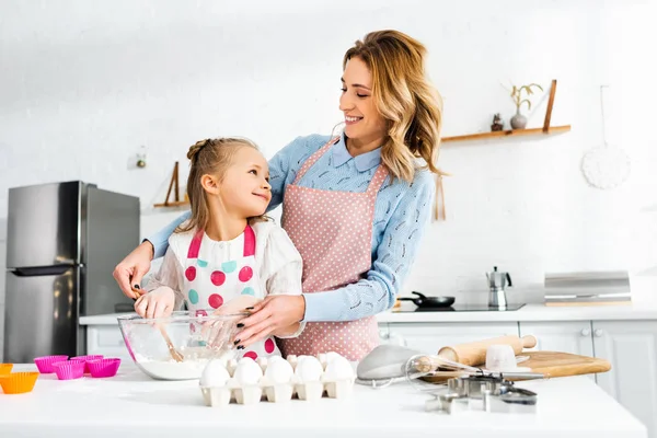 Hermosa mamá atractiva y linda hija mirándose y cocinando deliciosos cupcakes juntos - foto de stock