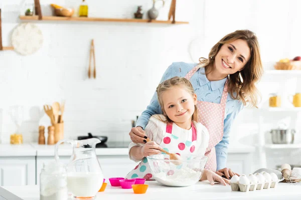 Enfoque selectivo de madre e hija sonriendo y cocinando deliciosos cupcakes con ingredientes como huevos, leche y harina - foto de stock