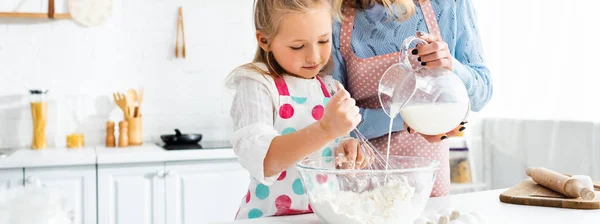 Figlia impastando la pasta mentre la madre versa il latte da brocca in boccia, colpo panoramico — Foto stock