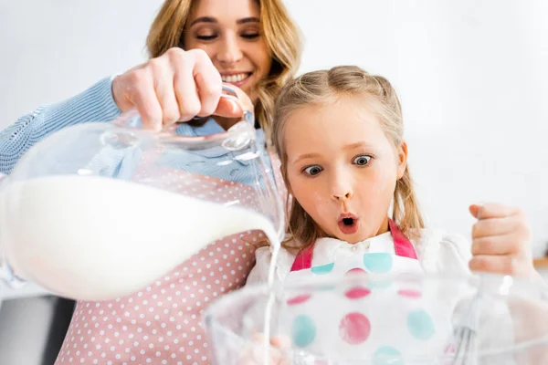 Vista de ángulo bajo de la hija conmocionada mirando a la madre vertiendo leche de la jarra en un tazón - foto de stock