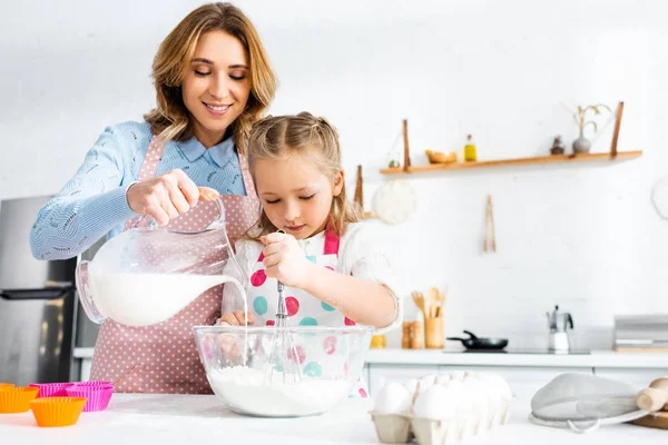 Hija amasando masa mientras la madre vierte la leche de la jarra en un tazón en la cocina - foto de stock