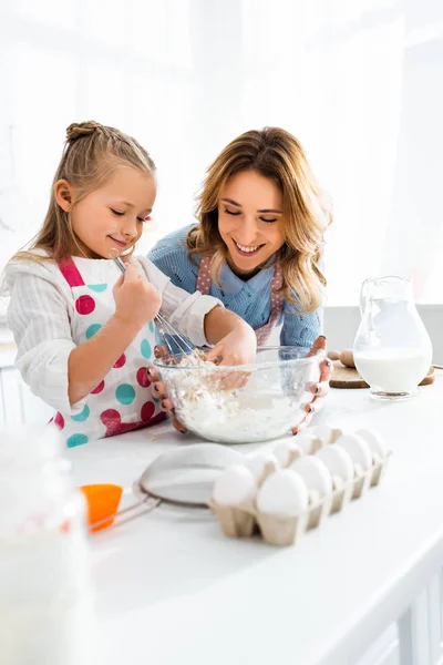 Focus selettivo della madre guardando figlia impastare pasta in ciotola a brocca di latte, uova, farina e utensili da cucina — Foto stock