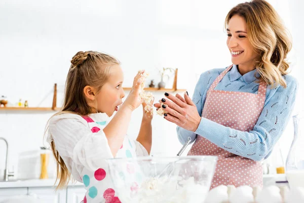 Selective focus of smiling mom and daughter with open mouth holding up hands having fun during kneading dough — Stock Photo