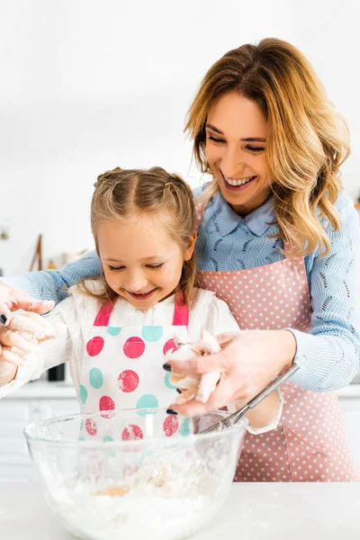 Селективный фокус привлекательной мамы и милой дочери улыбаясь и смешивая ингредиенты для вкусных кексов вместе — стоковое фото