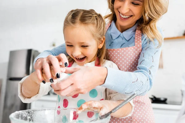 Concentration sélective de la mère attrayante et fille mignonne riant pendant l'ajout d'ingrédients à la pâte dans le bol — Photo de stock