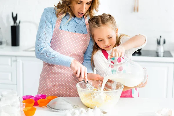 Vista ritagliata della mamma che cucina con frusta palloncino e figlia versando latte — Foto stock