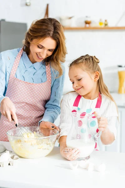 Sonriente madre cocinando con globo batidor e hija sosteniendo jarra con leche - foto de stock