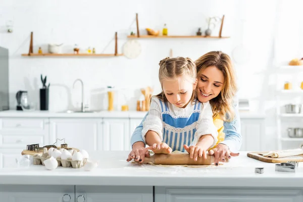 Sonriente madre y linda hija rodando masa en la mesa - foto de stock