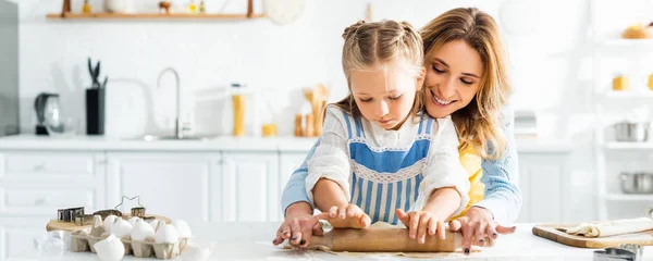 Panoramic shot of smiling mother and cute daughter rolling dough on table — Stock Photo