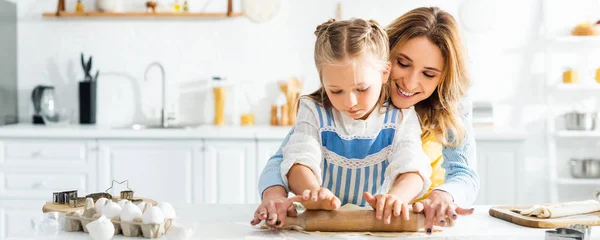 Colpo panoramico di madre sorridente e carina figlia rotolamento pasta sul tavolo — Foto stock