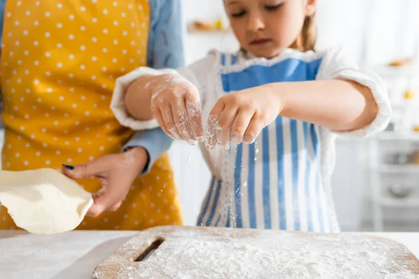 Vista recortada de la madre sosteniendo la masa y la hija tamizando la harina en la tabla de cortar - foto de stock