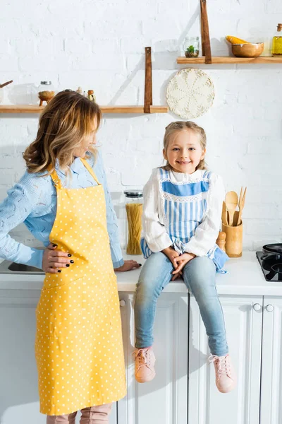 Mutter in Schürze schaut lächelnde und süße Tochter in der Küche an — Stockfoto