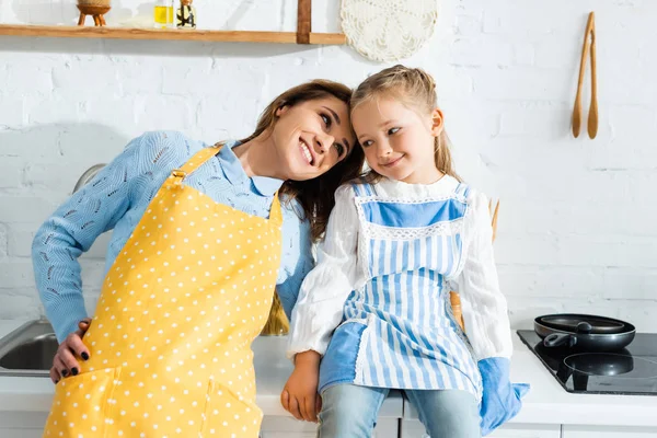 Smiling mother in apron hugging cute daughter in kitchen — Stock Photo