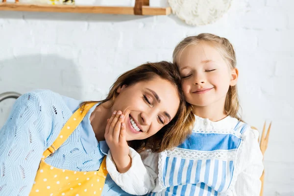 Sorrindo filha com olhos fechados abraçando mãe na cozinha — Fotografia de Stock
