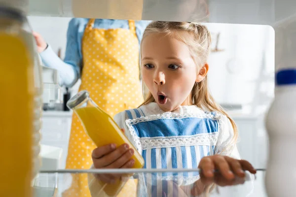 Messa a fuoco selettiva del bambino scioccato cercando e tenendo bottiglia con succo d'arancia — Foto stock