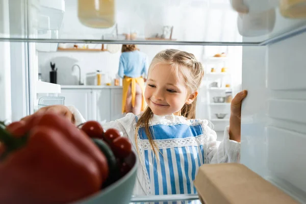 Foco seletivo de sorrir filha olhando para legumes na geladeira — Fotografia de Stock