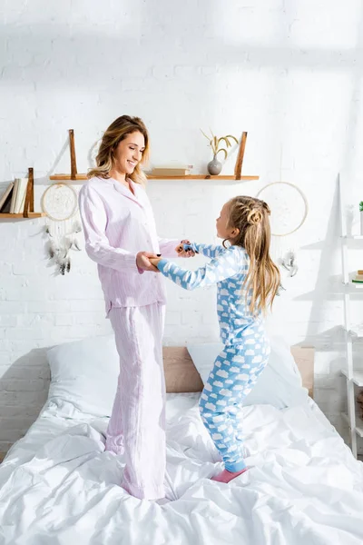 Smiling mother and daughter holding hands and jumping on bed — Stock Photo