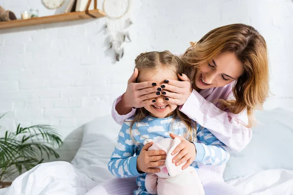 Sonriente y atractiva madre oscureciendo la cara de hija en pijama - foto de stock