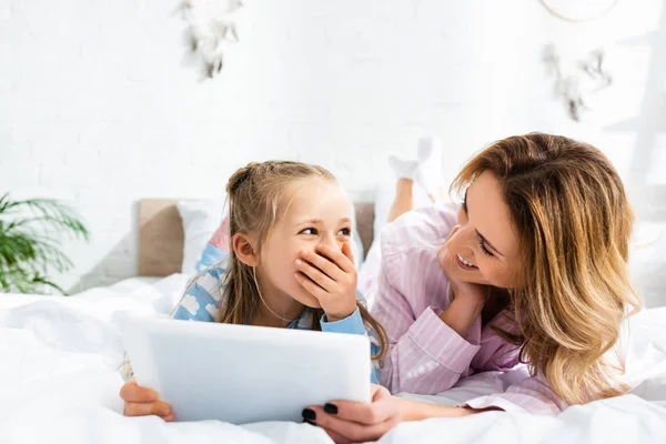 Lächelnde Mutter blickt geschockte Tochter mit digitalem Tablet im Schlafzimmer an — Stockfoto