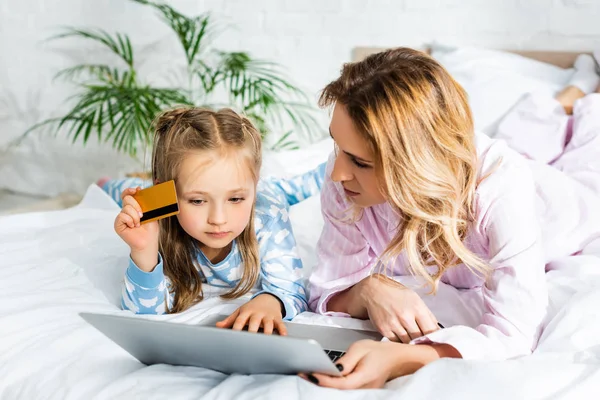 Madre mirando a la computadora portátil y la hija con tarjeta de crédito en el dormitorio - foto de stock